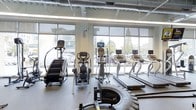 This fitness center offers a well-equipped, contemporary workout space designed to support an active lifestyle. The facility is lined with expansive floor-to-ceiling windows that flood the interior with abundant natural light while providing open views of the surrounding outdoor environment, creating an energizing atmosphere for exercise. The large glass panels contribute to a bright, airy feel that makes the space feel open and inviting.

The cardio area is outfitted with a comprehensive selection of equipment, including treadmills, elliptical machines, a stair climber, and a stationary bike, all featuring integrated digital display screens for tracking workout metrics. A tire training station adds a functional fitness element to the space. The equipment is arranged to maximize floor space and ensure ease of movement throughout the facility.

The ceiling features exposed industrial-style ductwork and ventilation systems, complementing the modern aesthetic while ensuring proper airflow throughout the space. The flooring consists of durable, textured rubber material suited for high-traffic fitness use. Together, these elements create a functional, well-lit, and thoughtfully designed fitness environment.