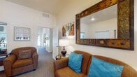This single-story clubhouse is designed to serve as a well-rounded community hub, offering multiple functional areas within a cohesive, inviting layout. The space features high ceilings throughout, lending an open and airy feel to each room.

The entry area greets visitors with carpeted floors, neutral walls, and tasteful decorative accents including colorful artwork and floral arrangements. Natural light filters in through windows near the entrance, creating a warm first impression. A pass-through window connects the lobby to an adjacent area, supporting community management and communications.

The lounge area is furnished with comfortable upholstered seating accented by vibrant teal throw pillows, complemented by a decorative oversized mirror and coordinating artwork. A water cooler and additional seating are available in a transitional lounge space that provides a view into the fitness room.

The business center is a dedicated, windowed room equipped with desktop computers, a printer, and built-in desk workspace, providing residents with convenient access to technology and productivity tools.

The fitness room is well-appointed with a multi-station weight machine, treadmill, climbing trainer, and stationary bike. Mirrored walls, a wall-mounted television, and natural light from large windows enhance the workout environment, making it both functional and motivating.