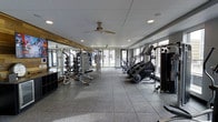This fitness center offers a well-appointed, single-level workout space designed to support a comprehensive exercise routine. The open floor plan provides ample room to move freely between stations, with durable rubber-tile flooring underfoot that ensures both safety and comfort during workouts. A ceiling fan mounted overhead complements the recessed lighting, contributing to a comfortable and well-ventilated environment. Large windows line the walls, flooding the space with natural light and offering views to the exterior, creating an open and energizing atmosphere. The strength training area is outfitted with a robust selection of free weights, weight racks, and multi-use lifting stations, all neatly organized along a mirrored wall that visually expands the space and allows exercisers to monitor their form. On the cardio side, the space features StairMaster machines and additional cardio equipment arranged to make the most of the available floor space. The combination of natural light, quality equipment, and a thoughtfully organized layout makes this fitness center a functional and inviting space suited for a wide range of fitness goals.