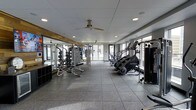 This fitness center offers a well-appointed, single-level workout space designed to support a comprehensive exercise routine. The open floor plan provides ample room to move freely between stations, with durable rubber-tile flooring underfoot that ensures both safety and comfort during workouts. A ceiling fan mounted overhead complements the recessed lighting, contributing to a comfortable and well-ventilated environment. Large windows line the walls, flooding the space with natural light and offering views to the exterior, creating an open and energizing atmosphere. The strength training area is outfitted with a robust selection of free weights, weight racks, and multi-use lifting stations, all neatly organized along a mirrored wall that visually expands the space and allows exercisers to monitor their form. On the cardio side, the space features StairMaster machines and additional cardio equipment arranged to make the most of the available floor space. The combination of natural light, quality equipment, and a thoughtfully organized layout makes this fitness center a functional and inviting space suited for a wide range of fitness goals.