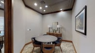 This single-floor space presents a sophisticated, intimate meeting room designed with careful attention to detail and material quality. The room is defined by warm walnut wood trim that frames the ceiling crown molding, baseboards, and the substantial entry doors, which feature glass panel inserts and open to a corridor showcasing exposed limestone masonry walls and wood beam accents beyond.

The ceiling is finished in a smooth neutral tone and equipped with recessed lighting that provides even, well-distributed illumination throughout the space. A central round table with a matte concrete top serves as the focal point, surrounded by mid-century modern-style chairs upholstered in charcoal fabric with curved walnut frames, comfortably seating a small group.

A patterned area rug in soft neutral tones anchors the seating arrangement and adds textural warmth to the floor. Along one wall, a geometric-front wooden sideboard displays a rectangular mirror flanked by two tall table lamps with white drum shades, contributing a polished, curated aesthetic. An abstract framed artwork in deep blues and golds is mounted on an adjacent wall, adding a refined artistic element. The overall ambiance is calm, professional, and thoughtfully appointed.