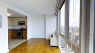 This high-rise residence offers a bright and open living environment defined by striking floor-to-ceiling windows that frame sweeping panoramic views of the city skyline and surrounding urban landscape. The generous windows flood the main living area with abundant natural light throughout the day, creating an airy and expansive atmosphere. Warm parquet hardwood flooring runs throughout the living space, adding texture and classic character to the contemporary setting. A perimeter heating unit is positioned along the window wall, ensuring year-round comfort while preserving the clean, open layout of the room. The kitchen is thoughtfully designed with modern cabinetry in a warm wood finish, stainless steel appliances including a gas range and microwave, and ample counter space — making it well-suited for everyday cooking. The open sightline between the kitchen and living area enhances the sense of connected, functional space. Crisp white walls provide a neutral canvas that complements both the natural wood tones and the urban views beyond. The overall ambiance is clean, modern, and filled with light, offering an elevated urban living experience with the dynamic cityscape as a constant backdrop.