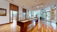 Located at 1203 Maritime Way in Ottawa, Ontario, this well-appointed amenity facility spans a generous single floor, offering a variety of recreational and social spaces designed for community engagement. The interior is anchored by warm, richly toned hardwood flooring that flows throughout the main areas, complemented by clean white walls and a drop ceiling fitted with adjustable track lighting that ensures bright, even illumination across the entire space.

The main room serves as a central social hub, featuring a full-length shuffleboard table as its focal point, alongside a upright piano and framed artwork that lend the area a refined yet welcoming ambiance. Adjacent to this is a dedicated ping-pong room, bathed in natural light from a series of large windows that offer views of mature trees and landscaped greenery outside.

A glass-walled room provides a more intimate setting, ideal for meetings or quiet gatherings, and is furnished with a dining table and chairs. Elegant wood-framed glass partitions and solid wood doors with warm walnut-toned finishes add architectural sophistication throughout. A smaller secondary room with light flooring and a full-length mirror appears suited for fitness or activity use. The entrance area features a curved glass facade, potted greenery, and display panels, creating an inviting and polished first impression.
