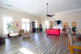 Located at 4129 Inverness Road in Virginia Beach, Virginia, this refined single-story community facility combines classic architectural detailing with functional, welcoming spaces throughout. The grand entrance foyer sets a distinguished tone, featuring rich hardwood-style flooring, detailed wainscoting and panel molding along the walls, built-in display niches, and a striking round center table crowned with a lush floral arrangement. Arched windows at the entry bathe the space in natural light. The expansive main lounge area is a standout, boasting soaring ceilings with crown molding, wide-plank hardwood-style floors, and an abundance of large windows that fill the room with daylight. A classic white fireplace serves as a focal point, accompanied by wall-mounted television for added comfort. Elegant panel molding continues throughout, reinforcing the space's refined aesthetic. Adjacent to the lounge, a dedicated business center offers a built-in workstation with multiple computer terminals, ergonomic seating, and a bright window overlooking greenery. The functional galley-style kitchen is equipped with ample cabinetry, granite-style countertops, tile flooring, a mosaic tile backsplash, and full-size black appliances, making it well-suited for catering and community events. Together, these spaces create a polished and inviting environment ideal for gatherings and everyday use.