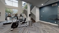 Located at 5005 Galleria Dr in Farmers Branch, TX, this two-story fitness center is a thoroughly appointed amenity space designed to support a wide range of workout routines. The facility is finished with stylish herringbone-patterned carpet throughout, crisp white walls accented by deep teal feature walls, and recessed lighting complemented by multiple ceiling fans for optimal air circulation.

The lower level serves as the main workout floor, housing a generous selection of Life Fitness strength machines, free weights, benches, stability balls, and cable systems. Large windows along one wall invite ample natural light and offer outdoor views, enhancing the energy of the space. A dedicated training zone near the entrance is equipped with a TRX suspension system, a punching bag, kettlebells, and a full accessory rack. An adjacent spin studio features stationary cycling bikes and a wall-mounted display screen for guided workouts.

A staircase with dark wood treads and a sleek metal railing connects the lower level to the upper floor, which continues the same design language and provides additional strength and cardio equipment. The upper level also features mirrored walls to amplify the sense of space. An elevator offers convenient access between floors. Together, both levels create a comprehensive, modern fitness environment suited for varied training needs.