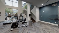 Located at 5005 Galleria Dr in Farmers Branch, TX, this two-story fitness center is a thoroughly appointed amenity space designed to support a wide range of workout routines. The facility is finished with stylish herringbone-patterned carpet throughout, crisp white walls accented by deep teal feature walls, and recessed lighting complemented by multiple ceiling fans for optimal air circulation.

The lower level serves as the main workout floor, housing a generous selection of Life Fitness strength machines, free weights, benches, stability balls, and cable systems. Large windows along one wall invite ample natural light and offer outdoor views, enhancing the energy of the space. A dedicated training zone near the entrance is equipped with a TRX suspension system, a punching bag, kettlebells, and a full accessory rack. An adjacent spin studio features stationary cycling bikes and a wall-mounted display screen for guided workouts.

A staircase with dark wood treads and a sleek metal railing connects the lower level to the upper floor, which continues the same design language and provides additional strength and cardio equipment. The upper level also features mirrored walls to amplify the sense of space. An elevator offers convenient access between floors. Together, both levels create a comprehensive, modern fitness environment suited for varied training needs.