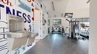 Located at 6655 Lookout Rd in Boulder, Colorado, this fitness center is a well-appointed exercise facility designed to support a full range of workout routines within a compact, efficiently organized footprint. The main workout area features durable rubber flooring throughout and is equipped with a variety of cardio machines, including treadmills and stationary spin bikes, alongside a multi-station strength training machine, free weights with a dedicated dumbbell rack, and an adjustable weight bench. A wall-mounted TRX suspension training system adds versatility for bodyweight exercises. Wall-mounted flat-screen televisions are positioned for easy viewing during workouts. The space is brightened by large windows along multiple walls, inviting generous natural light and offering outdoor views of the surrounding Boulder community. Vaulted ceilings with exposed ductwork contribute to an open, industrial-modern aesthetic. A bold, colorful typographic mural featuring fitness-themed words and icons energizes the space and serves as a striking visual focal point. A water fountain is conveniently accessible near the entrance. The facility also includes a clean, ADA-accessible restroom with tile flooring, a wall-mounted sink, and grab bars. The overall ambiance is motivating and functional, purpose-built to serve as a community fitness amenity.