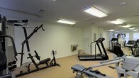 This single-floor fitness room offers a practical and functional workout space designed to accommodate a range of exercise routines. The room features white walls and a neutral-toned carpet floor, creating a clean and straightforward environment. Fluorescent ceiling fixtures provide consistent, even lighting throughout the space, ensuring good visibility during workouts.

The room is equipped with multiple pieces of exercise equipment, including at least two treadmills positioned along one side, a multi-station cable weight machine with an adjustable bench, and an additional workout bench with attachments for targeted strength training. A wall-mounted mirror on one side enhances the sense of space and allows users to monitor their form during exercise.

A built-in water fountain is conveniently located along one wall, providing easy hydration access. A small waste bin is positioned nearby. Ventilation is supported by ceiling vents distributed across the room. A door leads to an adjacent area, and a bulletin board is mounted near the exit. The overall ambiance is utilitarian and purposeful, catering to everyday fitness needs.