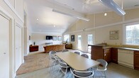 This single-floor community space is designed to accommodate both casual gatherings and relaxed lounging in a welcoming, open-concept layout. The room's most striking architectural feature is its vaulted ceiling, which rises dramatically to create an airy, expansive atmosphere enhanced by ceiling fans that promote comfortable airflow throughout. The interior is finished in warm neutral tones, with cream-colored walls and tile flooring that lend a clean, cohesive aesthetic. A central dining table surrounded by grey chairs provides a functional area for meetings or shared meals, while a dedicated lounge zone equipped with comfortable sofas and a wall-mounted flat-screen television offers a relaxed setting for residents or visitors. A built-in counter or bar area adds a practical service element to the space, ideal for hosting events or casual use. Natural light flows generously through multiple windows and glass-paneled doors, connecting the interior to an adjacent outdoor area visible through the entryways. Recessed lighting and pendant fixtures supplement the natural light, ensuring the space remains bright and inviting at any time of day. Overall, this community room balances functionality with comfort, making it well-suited for a variety of social and recreational activities.