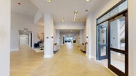 This expansive single-story amenity center serves as the social and functional hub of a modern multi-story residential community. The building's exterior features a striking combination of stone cladding and contemporary siding, complemented by a well-landscaped courtyard with lounge chairs, mature trees, and a pergola-covered gathering area.

Inside, the grand lobby welcomes visitors with light herringbone-patterned tile floors, white structural columns, recessed and accent lighting, and warm wood console tables. The soaring ceilings carry throughout, creating an airy, open atmosphere. The leasing office is appointed with a marble-topped reception desk, rich wood paneling, light hardwood-style flooring, and an eye-catching geometric accent wall. Private offices with glass partitions offer consultation spaces alongside a communal table for residents.

A social lounge area features a bold hexagonal tile backsplash, a long white quartz island with leather bar seating, pendant lighting, and a mounted television — ideal for casual gatherings. Wide corridors connect the various spaces, with light tile flooring, clean white walls, and a water fountain station.

The fitness center is well-equipped with a full array of cardio and strength machines, ceiling fans, wall-to-wall mirrors, and high clerestory windows that draw in ample natural light from multiple sides.