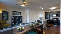 This single-floor residence showcases a thoughtfully designed open-concept layout that integrates the dining, living, and kitchen spaces into one cohesive and inviting environment. The dining area is anchored by a substantial dark wood table complemented by upholstered chairs in rich teal and neutral tones, creating a striking visual contrast. Decorative bowls of green fruit add a fresh, natural touch to the tablescape. A ceiling fan with a built-in light fixture overhead provides both comfort and ambient lighting, while recessed lighting throughout the space adds a clean, modern feel. Adjacent to the dining area, a dark bookcase styled with teal accents and white decorative objects serves as a bold design statement along the living space wall. The kitchen area is visible beyond, featuring a raised breakfast bar with contemporary bar stools, dark cabinetry, and light countertops that balance the overall color palette. Natural light filters in through a window near the kitchen, brightening the space. Neutral walls and warm hardwood-style flooring unify the entire open floor plan, while interior doors with classic paneling lead to additional rooms, rounding out this well-appointed and stylishly decorated home.