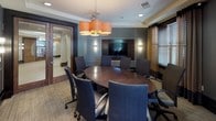 This professional conference room is thoughtfully designed to support productive meetings in a refined setting. At its center sits a substantial oval conference table with a rich, dark wood finish and high-gloss surface, surrounded by eight ergonomic upholstered chairs in a deep blue-gray fabric. A striking cluster of cylindrical pendant lights in warm amber tones serves as a focal point overhead, casting inviting light across the table.

The room features two distinct focal walls: one with a large mounted whiteboard framed by gray paneling with built-in sconce lighting, and an opposing wall housing a flat-screen television set within a dark wood built-in unit. Walls are finished in a sophisticated gray paneling treatment with contrasting trim, contributing to the room's executive character.

Natural light enters through several double-hung windows dressed with layered horizontal blinds and floor-length gold and charcoal drape panels, offering views of greenery outside. Textured loop-pile carpeting in neutral tones covers the floor. A glass-paneled door with dark wood framing provides access to an adjacent corridor, while recessed ceiling lighting supplements the pendant fixture for even, well-balanced illumination throughout.