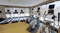 This fitness center is designed to provide a functional and comfortable workout environment. The space is lined with floor-to-ceiling windows along two walls, allowing abundant natural light to fill the room while offering views of the surrounding exterior. Roller shades are installed on the windows, giving users control over light and privacy during workouts. The cardio equipment selection is a notable highlight, featuring multiple treadmills equipped with individual display screens, as well as elliptical cross-trainers of varying styles and resistance levels, also fitted with personal monitors. A stair climber is positioned along the windowed wall, rounding out the cardiovascular offerings. Each machine is placed on protective rubber matting to preserve the flooring and reduce noise. The flooring itself features a patterned carpet in warm yellow and neutral tones, contributing to the space's inviting atmosphere. Recessed lighting in the ceiling supplements the natural light, ensuring consistent illumination throughout. Crown molding detail near the ceiling adds a polished, refined finish to the overall design of this practical and well-appointed fitness space.