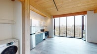 Located at 7373 N Philadelphia Ave in Portland, Oregon, this thoughtfully designed single-floor unit showcases a contemporary aesthetic rooted in natural materials and modern construction. The most striking architectural feature is the warm, exposed wood plank ceiling supported by timber beams, lending the space an inviting, organic character throughout.

The open living area is anchored by expansive floor-to-ceiling windows that frame sweeping views of the surrounding Portland neighborhood, flooding the interior with natural light and creating a strong sense of openness and connection to the outdoors. Light gray luxury vinyl plank flooring runs continuously through the main living spaces, complementing the warm tones of the wood ceiling above.

The kitchen is efficiently laid out along a single wall and is equipped with full stainless steel appliances, including a refrigerator, electric range, and dishwasher. Two-tone cabinetry — slate gray lower cabinets paired with white upper shelving — adds visual depth, while the clean countertops and under-cabinet range hood complete the functional design.

The bedroom benefits from the same dramatic corner window configuration, a wall-mounted mini-split HVAC unit, and the signature wood ceiling detail. The bathroom features a full tub-shower combo with a curved curtain rod, wood-toned flooring, and a dark-cabinetry vanity, offering a clean and practical layout. Overall, the space presents a cohesive, modern design with quality finishes throughout.