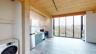 Located at 7373 N Philadelphia Ave in Portland, Oregon, this thoughtfully designed single-floor unit showcases a contemporary aesthetic rooted in natural materials and modern construction. The most striking architectural feature is the warm, exposed wood plank ceiling supported by timber beams, lending the space an inviting, organic character throughout.

The open living area is anchored by expansive floor-to-ceiling windows that frame sweeping views of the surrounding Portland neighborhood, flooding the interior with natural light and creating a strong sense of openness and connection to the outdoors. Light gray luxury vinyl plank flooring runs continuously through the main living spaces, complementing the warm tones of the wood ceiling above.

The kitchen is efficiently laid out along a single wall and is equipped with full stainless steel appliances, including a refrigerator, electric range, and dishwasher. Two-tone cabinetry — slate gray lower cabinets paired with white upper shelving — adds visual depth, while the clean countertops and under-cabinet range hood complete the functional design.

The bedroom benefits from the same dramatic corner window configuration, a wall-mounted mini-split HVAC unit, and the signature wood ceiling detail. The bathroom features a full tub-shower combo with a curved curtain rod, wood-toned flooring, and a dark-cabinetry vanity, offering a clean and practical layout. Overall, the space presents a cohesive, modern design with quality finishes throughout.