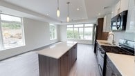 Unit 602 is a well-appointed 1-bedroom, 1-bathroom residence designed with a clean, contemporary aesthetic throughout its single floor. The open-concept living and kitchen area serves as the heart of the unit, creating a seamless flow between cooking and relaxing spaces. The kitchen is outfitted with dark wood cabinetry, a sleek quartz-topped center island with pendant lighting overhead, and stainless steel appliances including a gas range and refrigerator. Light wood-toned flooring runs throughout, complementing the neutral color palette of the walls and ceilings. A tray ceiling detail in the living area adds architectural depth and visual interest. Large, multi-pane windows along the living area wall flood the space with abundant natural light while framing elevated views of lush treetops and the surrounding neighborhood below — a standout feature of this sixth-floor unit. Recessed lighting throughout ensures the space remains bright and well-lit at all times. The overall ambiance is modern and airy, making excellent use of the open layout to create a comfortable and functional living environment suitable for a variety of lifestyles.