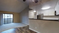 This single-floor unit showcases an open-concept design that integrates the living area and kitchen into one cohesive space. The vaulted ceilings create a sense of volume and airiness, enhanced by a ceiling fan that serves both the living and kitchen zones. The flooring transitions from rich, dark hardwood in the kitchen and entry areas to plush carpet in the living space, clearly defining each zone while maintaining visual continuity.

The kitchen features a peninsula with a dark countertop that doubles as a breakfast bar, providing both additional prep space and a casual dining option. White upper cabinetry offers clean contrast against the neutral gray walls, while a stainless steel refrigerator adds a modern, polished touch. A flush-mount light fixture illuminates the kitchen efficiently.

The living area is generously proportioned, with neutral carpeting and soft gray walls that create a calm, versatile backdrop. A doorway visible in the background hints at additional rooms beyond. The overall palette of warm neutrals, modern finishes, and practical layout make this a functional and inviting space.