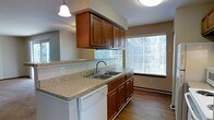 This single-story residence offers a well-designed layout that balances comfort and functionality. The kitchen is a standout feature, showcasing warm, rich-toned wood cabinetry that extends to the ceiling, providing ample storage space. The countertops display a granite-style finish, lending a clean and durable surface for meal preparation. A double-basin stainless steel sink sits beneath a window fitted with horizontal blinds, allowing for natural light to filter in while maintaining privacy. A pendant light fixture hangs above the counter peninsula, adding focused illumination and a touch of character to the space. The kitchen also includes a white dishwasher integrated seamlessly beneath the counter. An overhead cabinet structure frames the kitchen's open connection to the adjacent living area, which benefits from large windows that flood the space with bright, natural light. The flooring throughout transitions from the kitchen into the living area with warm hardwood-style planks, contributing to a cohesive and inviting atmosphere. The neutral wall tones complement the cabinetry and flooring, creating a balanced and welcoming interior environment throughout this thoughtfully arranged single-floor residence.