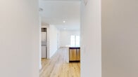 Located at 1564 Hi Point St in Los Angeles, this single-floor unit showcases a clean, modern design characterized by crisp white walls, recessed lighting, and light-toned hardwood flooring that runs consistently throughout the space. The open-concept layout connects the living area to a well-appointed kitchen, which features flat-front cabinetry in a warm natural wood finish, stainless steel appliances including a gas range and refrigerator, a vented range hood, and a distinctive blue patterned tile backsplash that adds a stylish focal point. A peninsula with a white countertop provides additional workspace and opens toward the living area, enhancing the sense of flow. A stacked in-unit washer and dryer is conveniently integrated into the layout. The bathroom is finished with a light wood vanity, white countertop, matte black fixtures, a framed mirror, and a tub-shower combination enclosed by sliding glass doors with black framing. Bedrooms are bright and airy, benefiting from large windows that frame outdoor views of the surrounding Los Angeles neighborhood. One room opens directly onto a private balcony through sliding glass doors, extending the living space outdoors and offering open views of the cityscape.