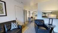This single-floor residence offers an open-concept layout that seamlessly connects the living, dining, and kitchen areas into one cohesive and functional living space. The living area is anchored by tufted leather seating, adding a classic touch to the neutral-toned interior. Light beige carpeting covers the main living and dining zones, while tile flooring marks the entryway near the front door, creating a natural transition between spaces.

The dining area sits centrally within the open layout, accommodating a modest table and chairs beneath an elegant chandelier that provides warm, focused lighting. Just beyond, the kitchen is partially visible, featuring white cabinetry and modern appliances, including what appears to be a stainless steel oven and range. A second pendant chandelier illuminates the kitchen area, contributing to the bright and welcoming atmosphere throughout.

The entryway includes a standard six-panel front door with an adjacent closet, providing convenient storage upon entry. The walls throughout are finished in a clean, neutral white, enhancing the sense of openness. Overall, the space presents a practical and well-organized layout suited for comfortable everyday living.