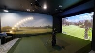 Located at 1200 Riverfront Dr in Little Rock, Arkansas, this 1,220-square-foot single-floor amenity space is divided into two distinct areas designed for entertainment and social gathering. The golf simulator room features dark, matte walls that create an immersive environment, anchored by a large-format Trackman projection screen, synthetic turf hitting surface, and dedicated club storage. Seating areas with plush armchairs and a window offering outdoor views add comfort alongside the high-tech setup. Golf-themed artwork and recessed lighting complete the room's focused aesthetic.

The adjoining lounge area presents a dramatic contrast, featuring exposed wood ceiling beams, dark structural columns, and light wood-look flooring. A large quartz-topped island with pendant lighting serves as the centerpiece, complemented by a full wet bar with a beverage cooler and coffee station. Geometric accent walls, upholstered banquette seating, and mixed dining and high-top tables provide flexible social spaces. A billiards table anchors one end of the room, while large windows introduce natural light, creating a polished yet relaxed atmosphere throughout.