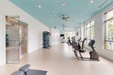 Located at 20013 Century Blvd in Germantown, MD, this well-appointed fitness center offers a generously sized single-floor layout designed to accommodate a wide range of exercise activities. The main exercise room is bright and airy, characterized by striking teal-painted ceilings accented with recessed lighting and multiple ceiling fans that keep the space comfortable. Large multi-pane windows line the walls, flooding the interior with abundant natural light and offering views of the surrounding greenery outside. The durable, light-colored flooring extends throughout, providing a clean and practical surface suited for all types of workouts. Equipment includes treadmills, elliptical trainers, stationary bikes, cable machines, a full dumbbell rack with benches, and functional training stations, all arranged to maximize usable floor space. A separate studio room features floor-to-ceiling mirrors, a ballet barre, and cushioned flooring, making it ideal for yoga, stretching, or group classes. A decorative floral mural adds an artistic touch to one section of the main floor. Additional amenities include a wall-mounted television, a water fountain station, and an accessible restroom. The overall ambiance is modern, clean, and energizing, creating an inviting environment for fitness enthusiasts of all levels.