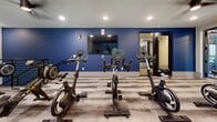 Located in Alpharetta, Georgia, this two-floor fitness center offers a comprehensive and well-appointed facility designed to support a wide range of workout routines. The ground floor serves as the primary training hub, housing an expansive open gym area with patterned carpet tile flooring, ceiling fans, and recessed lighting. This level features a robust selection of strength training equipment, including cable machines, weight benches, dumbbell racks, and multi-use resistance stations, all complemented by full-length mirrors along the walls. A dedicated cardio zone on this floor includes treadmills and stationary machines, bathed in natural light from elevated clerestory windows. A cycling studio features bold blue accent walls, wall-mounted flat-screen TVs, and a full row of stationary bikes on wood-look flooring. The upper floor continues the fitness-focused layout with an additional cycling area featuring large windows, ceiling fans, and hardwood-style flooring with open railing views below. A bright studio room on this level is ideal for yoga, barre, or group fitness classes, boasting floor-to-ceiling mirrors, balance balls, resistance equipment, and direct access to an outdoor balcony with abundant natural light. The facility maintains a clean, modern aesthetic throughout with a neutral color palette accented by navy blue feature walls.