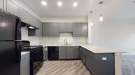 This single-floor unit offers a thoughtfully designed layout with contemporary finishes throughout. The kitchen serves as a standout feature, showcasing gray shaker-style cabinetry that extends to the ceiling, providing generous storage. Marble-look countertops line the L-shaped workspace, paired with a stainless steel sink and coordinating hardware. Black appliances — including a gas range, dishwasher, and full-size refrigerator — add a bold contrast to the light color palette. Pendant lighting above the peninsula and recessed ceiling lights illuminate the space effectively. Light wood-look luxury vinyl plank flooring runs throughout the main areas, lending warmth and easy maintenance. The bedroom features neutral carpet and a window overlooking an outdoor balcony area, allowing natural light to filter in. The bathroom is notably spacious and accessibility-ready, equipped with grab bars around the tub-shower combination, a comfort-height toilet, and a gray vanity with a marble-look countertop that mirrors the kitchen's design. The living area provides a generous open footprint with a ceiling fan, connecting seamlessly to the rest of the unit. Clean white walls and recessed lighting throughout maintain a bright, cohesive atmosphere across all rooms.