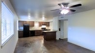 This single-story space showcases an open-concept layout that seamlessly connects the kitchen and living areas. The polished concrete floors extend throughout, providing a durable and modern foundation that complements the overall design. The kitchen is well-appointed with rich, dark wood cabinetry offering generous storage space, and black appliances including a refrigerator, range, and dishwasher. A peninsula-style island with a light-toned countertop serves as both a prep area and a casual dining or gathering spot, anchoring the kitchen within the open floor plan. Recessed lighting and flush-mount ceiling fixtures provide consistent illumination throughout the space. The living area features a ceiling fan with a built-in light kit, contributing to both comfort and functionality. A hallway visible from the main living space leads to additional areas of the unit, including what appears to be a closet with bifold doors. The neutral white walls contrast cleanly with the darker flooring and cabinetry, creating a balanced, practical interior. The open layout and polished concrete floors lend the space an industrial yet inviting character.