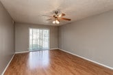This single-floor residence is characterized by consistent warm-toned hardwood-style flooring that runs throughout the main living areas and bedrooms, tying the space together cohesively. The layout, as revealed by the 3D overview, features a connected living and dining area that flows naturally toward the kitchen, creating an open, functional arrangement.

The living area is generously sized and benefits from a ceiling fan with a light fixture, neutral gray walls, and a sliding glass door that allows natural light to fill the room while providing outdoor access. The textured ceilings are present throughout the unit.

The kitchen is efficiently designed with warm wood-toned cabinetry, a white refrigerator, and a cooktop range. A raised breakfast bar with an overhanging counter separates the kitchen from the living space, offering a casual dining or workspace option.

The bedrooms feature the same hardwood-style flooring, neutral gray walls, and vertical blind-covered windows. One bedroom includes a sliding closet door, providing practical storage. An additional room with a window and closet offers flexible use.

The bathroom is clean and functional, featuring a tiled tub-and-shower combination, a single-sink vanity with wood cabinetry, a framed mirror, and tile flooring.