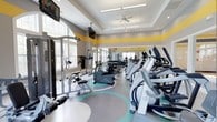 This fitness center is designed to support a comprehensive workout experience in a spacious, thoughtfully appointed environment. The interior is bright and welcoming, with large windows along the perimeter allowing abundant natural light to fill the space. A tray ceiling with recessed lighting and a ceiling fan adds architectural interest while maintaining excellent ventilation and visibility throughout. Bold yellow accent stripes along the upper walls provide an energizing pop of color that complements the neutral gray tones of the walls and the decorative, multi-toned flooring below — which features sweeping circular patterns in teal and white, adding a dynamic visual element underfoot. The facility is well-stocked with a full row of cardio machines, including ellipticals and stationary bikes, positioned to take advantage of the natural light. A cable and multi-function strength training station anchors one side of the room, while a dumbbell rack and additional strength equipment round out the offerings. Wall-mounted televisions are positioned throughout for entertainment during workouts. Full-length mirrors along one wall enhance the sense of openness and allow users to monitor their form. The overall ambiance is clean, modern, and motivating — a fully functional space suited for a wide range of fitness routines.