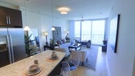 This single-floor space offers an open-concept layout that combines the dining and living areas into one cohesive, well-appointed environment. The dining area features a granite-topped counter or bar with wire-frame chairs, providing a stylish and functional space for casual meals. Place settings and natural woven placemats add a refined yet relaxed touch to the surface.

The living area is generously sized, anchored by a plush sofa adorned with decorative pillows and complemented by a pair of patterned accent chairs arranged around a dark wood coffee table. A large area rug defines the seating zone, while warm ambient lighting from floor lamps and a central ceiling fixture creates an inviting atmosphere.

The most striking feature is the expansive floor-to-ceiling window wall, which floods the interior with natural light and offers panoramic views of the outdoors, including what appears to be an open sky and balcony access. Light, neutral wall tones and light hardwood-style flooring enhance the airy, coastal feel of the space, making it feel open and serene throughout.