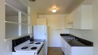 This single-floor space includes a compact, efficiently laid out kitchen that maximizes its available footprint. The kitchen features white cabinetry along both walls, providing ample storage through upper and lower cabinets, as well as open shelving in one corner. Dark granite-style countertops contrast cleanly against the white cabinetry, lending a classic aesthetic to the space. A double-basin sink is positioned along one wall, accompanied by standard fixtures. The kitchen is equipped with a white coil-burner electric range and a full-size white refrigerator, offering all essential appliances. A flush-mount ceiling light provides overhead illumination, keeping the space bright and functional. The light-colored walls contribute to an open, airy feel despite the kitchen's modest scale. Neutral toned flooring runs throughout, complementing the overall color palette. The layout is designed as a galley-style kitchen, with counters and appliances arranged along parallel walls to create a practical and organized workflow. Overall, the space presents a clean, straightforward design suited for everyday living.