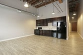 This distinctive loft-style space showcases a compelling blend of industrial heritage and contemporary design across a single floor. The open-concept living and kitchen area commands attention with soaring ceilings supported by exposed wooden beams and visible structural elements, lending the space an authentic industrial character. Light wood-look vinyl plank flooring runs throughout the main living areas, complementing the warm tones of the exposed brick accent wall, which features a window that allows natural light to filter in.

The kitchen is well-appointed with rich dark espresso cabinetry offering ample storage, light countertops, and a full suite of black appliances including a refrigerator with ice maker, range, built-in microwave, and dishwasher. Industrial-style pendant lighting illuminates the kitchen and living areas, reinforcing the loft aesthetic.

A carpeted room provides a quieter, softer-underfoot space suitable for a bedroom or private retreat, with exposed ductwork along the ceiling maintaining the industrial theme. The bathroom is clean and functional, featuring a dark vanity with a striped stone-look countertop, a white under-mount sink, recessed lighting, and a full bathtub and shower combination with tile flooring. Together, these elements create a cohesive, urban loft atmosphere that is both stylish and practical.