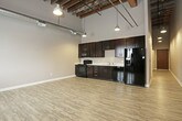 This distinctive loft-style space showcases a compelling blend of industrial heritage and contemporary design across a single floor. The open-concept living and kitchen area commands attention with soaring ceilings supported by exposed wooden beams and visible structural elements, lending the space an authentic industrial character. Light wood-look vinyl plank flooring runs throughout the main living areas, complementing the warm tones of the exposed brick accent wall, which features a window that allows natural light to filter in.

The kitchen is well-appointed with rich dark espresso cabinetry offering ample storage, light countertops, and a full suite of black appliances including a refrigerator with ice maker, range, built-in microwave, and dishwasher. Industrial-style pendant lighting illuminates the kitchen and living areas, reinforcing the loft aesthetic.

A carpeted room provides a quieter, softer-underfoot space suitable for a bedroom or private retreat, with exposed ductwork along the ceiling maintaining the industrial theme. The bathroom is clean and functional, featuring a dark vanity with a striped stone-look countertop, a white under-mount sink, recessed lighting, and a full bathtub and shower combination with tile flooring. Together, these elements create a cohesive, urban loft atmosphere that is both stylish and practical.