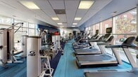Located at 222 E Bland St in Charlotte's South End neighborhood, this single-floor fitness center is designed to accommodate a comprehensive range of workout needs. The space is defined by its bold blue accent walls and vibrant blue carpet flooring, creating an energetic and motivating atmosphere throughout.

Along one side of the facility, a row of cardio machines — including treadmills and ellipticals equipped with individual screens — lines the expansive floor-to-ceiling windows, flooding the space with natural light and providing views of the surrounding urban environment. The layout maximizes usable floor space while maintaining an open, accessible feel.

The strength training section is well-appointed with a variety of Cybex cable machines, Smith machines, weight benches, and plate-loaded equipment, offering users a full range of resistance training options. A dedicated free weights area features a tiered dumbbell rack alongside a wall-mounted flat-screen TV, providing both convenience and entertainment during workouts. Stability balls and yoga mats are also available for functional training and stretching.

Overhead, a drop ceiling with recessed fluorescent panel lighting ensures consistent, even illumination throughout the facility. The space is equipped with sanitation stations to support cleanliness and user safety. Overall, the fitness center offers a well-organized, fully equipped environment suitable for a variety of fitness routines.