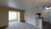 This single-story space in Irving, Texas, presents an open and functional layout that connects the main living area to the kitchen and beyond. The living space is finished with wall-to-wall carpet, offering a comfortable and warm underfoot feel throughout. A textured popcorn ceiling adds dimension overhead, while multiple HVAC vents indicate a well-distributed climate control system.

A wide, arched passageway leads from the main living area into a secondary space illuminated by a large window fitted with horizontal blinds, allowing for adjustable natural light. A ceiling fan fixture in that adjoining area enhances airflow and provides additional overhead lighting.

To one side of the living area, a built-in shelving unit offers convenient open storage, seamlessly transitioning into the kitchen space. The kitchen features a dark-toned countertop with a sink, dark tile backsplash, and a glimpse of appliances, suggesting a compact yet practical cooking area. Neutral beige walls throughout the space create a clean, understated aesthetic that lends itself well to a variety of interior styles.