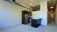 This single-floor space showcases a contemporary industrial aesthetic, highlighted by soaring ceilings with exposed silver ductwork and track lighting that runs across the ceiling, lending a distinctly urban loft character. The open-plan layout seamlessly integrates the kitchen and living areas into one cohesive space.

The kitchen is a standout feature, outfitted with dark espresso cabinetry that contrasts sharply against the light-toned walls and flooring. Stainless steel appliances — including a refrigerator, microwave, and range — provide a sleek, modern finish. A peninsula with a granite-style countertop and an integrated sink extends into the living area, offering additional prep space and a natural room divider that maintains the open flow of the layout.

Light carpet covers the living area floor, offering a soft contrast to the kitchen's harder surfaces. The clean, minimalist walls reflect ample light from the directional track fixtures overhead. The combination of industrial design elements with modern finishes creates a functional yet stylish atmosphere well-suited to contemporary urban living.