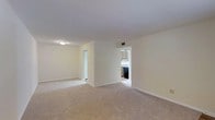 This single-floor residential space presents a clean, straightforward layout designed for comfortable everyday living. The main living area is generously sized, featuring wall-to-wall carpeting in a neutral beige tone that adds warmth underfoot and complements the soft, cream-colored walls throughout. A textured ceiling with flush-mount lighting provides even illumination across the open room, while a visible HVAC vent ensures year-round climate comfort. The living space connects seamlessly to additional areas via open doorways, promoting an easy flow throughout the home. One passage leads to what appears to be a secondary room or hallway, offering flexible use of space. A second opening provides direct sightlines into the kitchen, which features wood-toned cabinetry, a dark appliance suite, and a sink positioned beneath upper cabinets — a practical and efficient cooking area. The overall aesthetic is neutral and move-in ready, with a consistent color palette of whites and beiges that creates a bright, airy atmosphere. The minimalist design allows for a wide range of furnishing and personalization options, making this space both versatile and functional.
