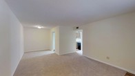 This single-floor residential space presents a clean, straightforward layout designed for comfortable everyday living. The main living area is generously sized, featuring wall-to-wall carpeting in a neutral beige tone that adds warmth underfoot and complements the soft, cream-colored walls throughout. A textured ceiling with flush-mount lighting provides even illumination across the open room, while a visible HVAC vent ensures year-round climate comfort. The living space connects seamlessly to additional areas via open doorways, promoting an easy flow throughout the home. One passage leads to what appears to be a secondary room or hallway, offering flexible use of space. A second opening provides direct sightlines into the kitchen, which features wood-toned cabinetry, a dark appliance suite, and a sink positioned beneath upper cabinets — a practical and efficient cooking area. The overall aesthetic is neutral and move-in ready, with a consistent color palette of whites and beiges that creates a bright, airy atmosphere. The minimalist design allows for a wide range of furnishing and personalization options, making this space both versatile and functional.