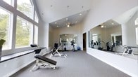 This single-floor fitness facility offers a bright, open, and welcoming environment designed to support a comprehensive workout experience. The space is defined by its impressive vaulted ceilings, which lend a sense of openness and airiness throughout. Large windows line one side of the room, flooding the interior with abundant natural light and providing views of the surrounding greenery outdoors. A ceiling fan is centrally mounted, ensuring comfortable air circulation during workouts.

The fitness area is carpeted and thoughtfully laid out with a variety of strength training equipment, including adjustable benches and weight machines positioned for ease of use. Cardio equipment is also present, rounding out the facility's offerings for a full-body workout. Large wall-mounted mirrors are positioned strategically throughout the space, reflecting light and allowing users to monitor their form during exercise. A stability ball and additional accessories complement the equipment selection.

The clean, neutral color palette of white walls and light grey carpet creates a calm and focused atmosphere. An adjoining doorway suggests additional access to connected spaces. Overall, this fitness facility combines functional design with an airy, well-lit ambiance, making it an inviting space for daily exercise and wellness routines.