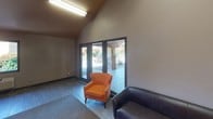 This single-floor communal space is thoughtfully designed to serve as both a relaxed lounge and a functional workspace. The room is defined by its striking vaulted ceilings, which lend a sense of openness and volume that extends well above the main living area. Warm, earthy tones dominate the interior — rich amber and tan walls create an inviting, grounded atmosphere throughout.

On one side of the space, a long built-in granite-topped desk runs the length of the wall, accompanied by patterned upholstered chairs with dark wood legs and integrated storage drawers beneath. This dedicated workspace is ideal for focused tasks and productivity. A wall-mounted HVAC unit and a square window framing a view of lush outdoor greenery add both comfort and natural light to this corner of the room.

The lounge area is furnished with a dark leather sofa and a vibrant orange accent chair, positioned to take full advantage of the room's most striking feature — a set of large glass doors that open directly to an outdoor patio area. The patio reveals stonework columns, a picnic area, and native landscaping, blurring the boundary between indoor and outdoor living. A pass-through window connects to an adjacent room, enhancing the space's versatility. Tile flooring and area rugs complete the functional, community-oriented design.