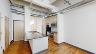 Located at 311 South 11th Street in Richmond, VA, this urban loft-style unit showcases a compelling blend of industrial and modern design across a single floor. The open-concept kitchen serves as a central focal point, featuring a granite-topped island with an undermount sink, stainless steel appliances including a gas range, microwave, and refrigerator, complemented by light maple cabinetry and a mosaic tile backsplash. Pendant lighting hangs from the exposed ceiling, where structural beams, track lighting, and metallic ductwork contribute to the space's distinctive industrial character. Rich hardwood floors run throughout the main living areas and bedrooms, adding warmth that contrasts beautifully with the raw, utilitarian ceiling elements. The bedroom areas feature painted brick walls and large windows that invite natural light and offer views of the surrounding urban streetscape. The bathroom is finished with light tile flooring, a full tub-shower combination with white subway tile surround, and a warm wood vanity topped with a dark countertop. Overall, the space delivers an authentic loft aesthetic with practical modern finishes, well-suited for urban living in the heart of Richmond.