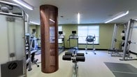 This fitness center offers a thoughtfully designed workout space finished with sage green walls, light-colored carpeting, and a clean white ceiling. A standout architectural feature is a tall, cylindrical column clad in a rich, warm-toned perforated metal casing, serving as a bold visual focal point at the center of the room. Pendant lighting runs along the ceiling, complementing the natural light that flows in through multiple large, frosted windows along the far wall, creating a bright and energizing atmosphere. The cardio section is well-appointed with several treadmills, each equipped with individual monitor screens for entertainment during workouts. An elliptical trainer and a stair climber round out the cardiovascular offerings. Strength and flexibility training is supported by a dedicated ab bench with padded rollers, positioned prominently in the foreground. A multi-function weight machine is also visible near the entrance. The layout is open and spacious, allowing for comfortable movement between equipment. Overall, the space balances functional fitness amenities with a clean, modern aesthetic suited for a range of workout routines.