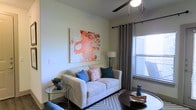 This single-floor space offers a comfortable and stylishly designed living environment. The living room serves as the focal point, showcasing a light-colored sofa accented with blue and patterned throw pillows, arranged around a dark circular coffee table. A large, bold abstract artwork in warm red and orange tones anchors the wall, adding a striking visual element to the otherwise neutral color palette. The room is finished with light-toned walls and smooth ceilings, with a ceiling fan providing both function and a casual aesthetic. Natural light filters in generously through a window dressed with gray grommet-panel curtains and horizontal blinds, creating a bright and airy atmosphere. A glass-topped side table, a floor lamp, and a textured area rug complete the thoughtfully curated space. The overall ambiance is modern yet inviting, blending clean lines with warm accents to create a space that feels both livable and aesthetically considered.