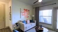 This single-floor space offers a comfortable and stylishly designed living environment. The living room serves as the focal point, showcasing a light-colored sofa accented with blue and patterned throw pillows, arranged around a dark circular coffee table. A large, bold abstract artwork in warm red and orange tones anchors the wall, adding a striking visual element to the otherwise neutral color palette. The room is finished with light-toned walls and smooth ceilings, with a ceiling fan providing both function and a casual aesthetic. Natural light filters in generously through a window dressed with gray grommet-panel curtains and horizontal blinds, creating a bright and airy atmosphere. A glass-topped side table, a floor lamp, and a textured area rug complete the thoughtfully curated space. The overall ambiance is modern yet inviting, blending clean lines with warm accents to create a space that feels both livable and aesthetically considered.