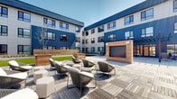 This expansive courtyard is set within a modern multi-story building clad in bold navy blue horizontal siding and crisp white paneling, creating a striking contemporary backdrop. The outdoor space is thoughtfully organized into several distinct functional zones, each designed to encourage both social interaction and relaxation. A central lounge area features a patterned deck of interlocking wood-look tiles, furnished with woven rattan chairs and cushioned ottomans arranged around a wood-clad outdoor fireplace, making it a natural gathering point. Adjacent to this is an outdoor kitchen station finished in warm cedar, equipped with a stainless steel grill and prep sink, paired with white dining tables and vibrant yellow chairs beneath a sleek pergola. Raised cedar planter beds introduce greenery and landscaping throughout, softening the hardscape and adding warmth to the modern aesthetic. A dedicated sun deck area offers chaise lounges set against manicured lawn panels and curated planting beds. A covered patio near the building entrance features a pergola with open-beam detailing and comfortable lounge seating, seamlessly connecting the indoors and outdoors through floor-to-ceiling glass doors. The overall ambiance is polished, welcoming, and designed for active community living.