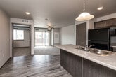 Located at 1360 NE Primrose Ln in Grimes, Iowa, this 1-bedroom, 1-bathroom residence offers a thoughtfully designed single-floor layout that maximizes functionality and comfort. The open-concept kitchen and living area serves as the heart of the home, where wood-look luxury vinyl flooring flows seamlessly underfoot. The kitchen features a peninsula with a double-basin sink and pendant lighting overhead, dark-toned cabinetry, a black refrigerator, and a black range with over-the-range microwave — all complementing the neutral countertops. The living area extends naturally from the kitchen, finished with plush carpeting and a ceiling fan, and opens directly onto a private balcony through sliding glass doors, bringing in natural light and outdoor access. A dedicated pantry provides additional storage. The bedroom is a comfortable, carpeted retreat with a window offering natural light, and is serviced by a well-sized closet. The bathroom includes a dark-cabineted vanity with a sink, mirror, and wood-look flooring. A dedicated laundry room houses a full-size washer and dryer with built-in wire shelving above. The textured ceilings and neutral color palette throughout create a clean, cohesive aesthetic.