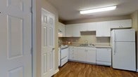 This single-floor residential space offers a clean, functional layout designed for comfortable everyday living. The kitchen stands out as a central feature, showcasing white shaker-style cabinetry that extends to the upper walls, providing generous storage space. The countertops offer ample workspace, and the kitchen is equipped with a white electric range, a built-in dishwasher, and a stainless steel sink with a modern faucet. An overhead range hood is integrated seamlessly into the cabinetry above the stove. Warm hardwood-style flooring runs throughout the kitchen, adding a touch of natural warmth that contrasts nicely with the bright white finishes. A textured ceiling with recessed fluorescent lighting ensures the space is well-lit. The six-panel interior door visible in the foreground is a classic architectural touch that complements the overall clean aesthetic. The neutral wall tones create a versatile backdrop, making the space easy to personalize. The layout flows efficiently, offering a practical and inviting environment suited for daily living.