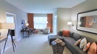 This well-appointed single-floor space showcases a harmonious blend of comfort and function. The living area serves as the centerpiece, featuring plush wall-to-wall carpeting in a soft neutral tone that complements the warm beige walls throughout. A generously sized sectional sofa in charcoal gray anchors the room, accented with vibrant rust-orange throw pillows that echo the rich, floor-length drapery framing the large windows. The windows bathe the space in natural light while offering a bright, welcoming ambiance. A tufted ottoman and a decorative round side table complete the seating arrangement, adding both style and practicality. Toward the far end of the room, a dedicated workspace features a sleek dark wood desk and chair, ideal for productivity. A patterned accent chair positioned near the window creates a cozy reading nook, perfectly placed to take advantage of the natural light. Wall-mounted and freestanding lamps provide layered lighting throughout the space. The overall design aesthetic is modern yet inviting, with a cohesive color palette of warm neutrals and burnt orange accents that lend the space a polished, curated feel.